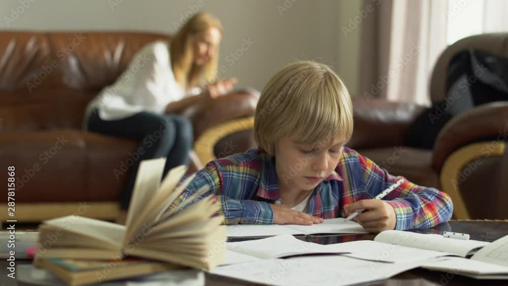 Cute little boy writing down information in the notebook at home in the foreground while his mother sitting in the background. Studying. Adorable schoolkid doing lessons. Cute little boy writing down information in the notebook at home in the foreground while his mother sitting in the background. Studying. Adorable schoolkid doing lessons.