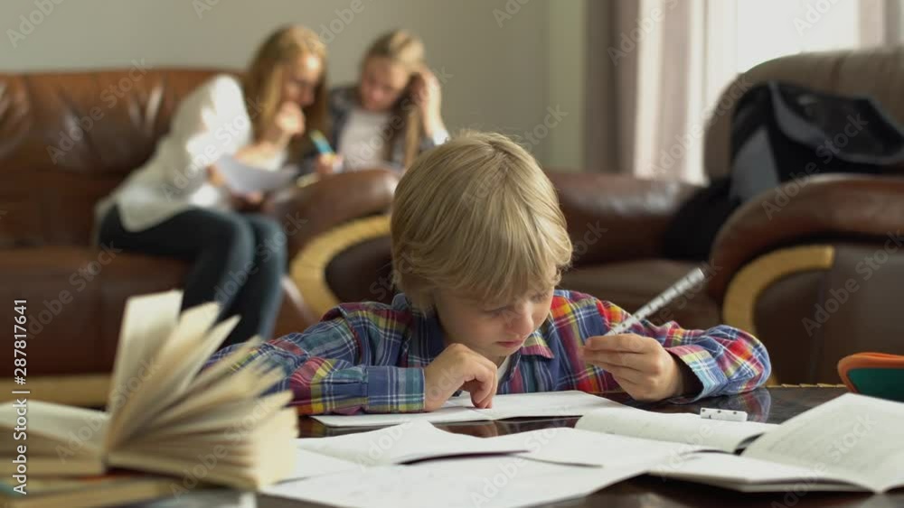 Little boy sitting at the table with notebooks at home while doing homework. Mother and sister sitting in the background looking pappers. Schoolboy rubs tired eyes with his hand. Little boy sitting at the table with notebooks at home while doing homework. Mother and sister sitting in the background looking pappers. Schoolboy rubs tired eyes with his hand.