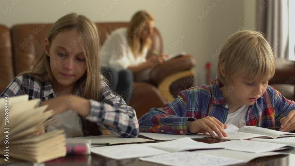 Adorable little boy and girl studying at home in the foreground while their mother sitting in the background. Schoolkids doing their homework together. Adorable little boy and girl studying at home in the foreground while their mother sitting in the background. Schoolkids doing their homework together.