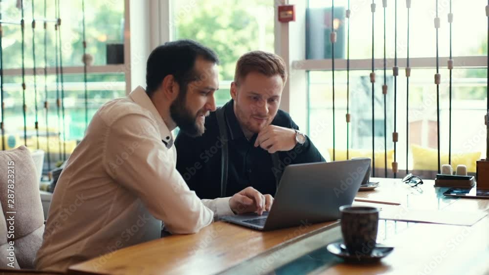Portrait of two young business guy sitting at table with laptop near the window in cozy cafe. Shot in slow motion Portrait of two young business guy sitting at table with laptop near the window in cozy cafe. Shot in slow motion