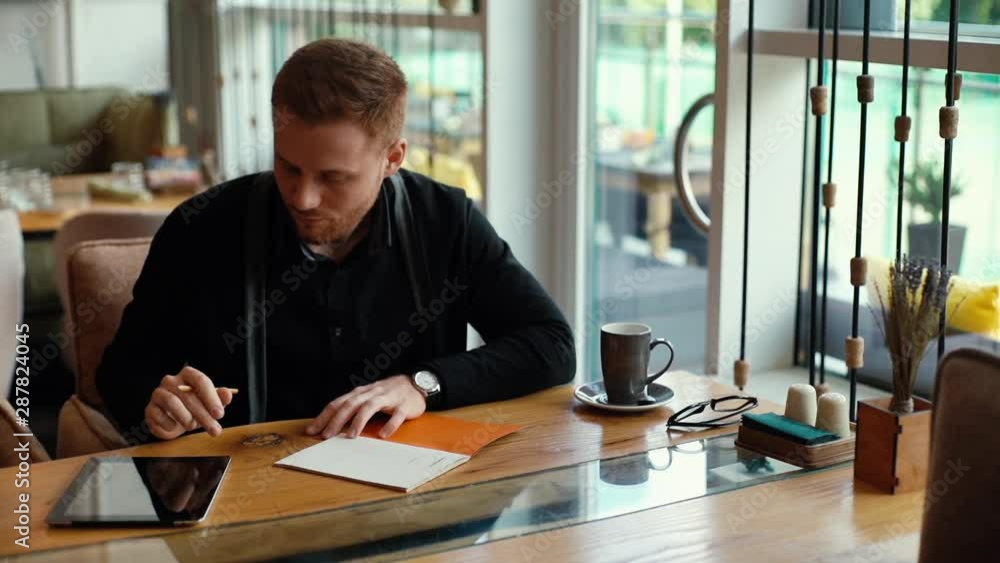 Young handsome man writes in notebook and works on tablet in cozy cafe interior. Attractive young businessman is writing in notepad. Shot in slow motion Young handsome man writes in notebook and works on tablet in cozy cafe interior. Attractive young businessman is writing in notepad. Shot in slow motion