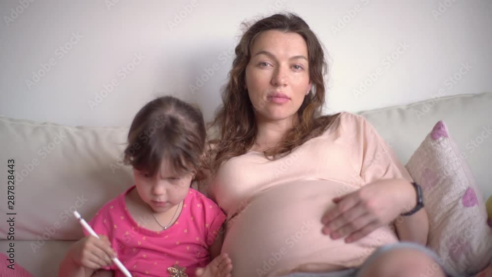 Young happy pregnant mother preparing an older child for a new baby, sitting on sofa in white interior. A woman teaches a girl drawing on a tablet computer with digital pen. Young happy pregnant mother preparing an older child for a new baby, sitting on sofa in white interior. A woman teaches a girl drawing on a tablet computer with digital pen.