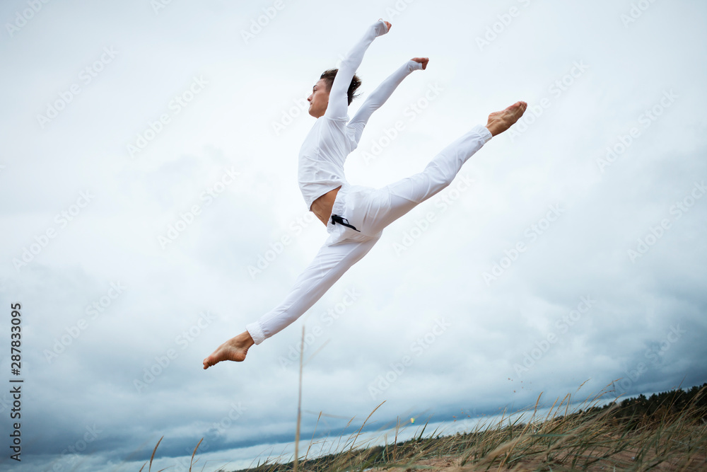Outdoor portrait of stylish flexible young Caucasian man in white sports outfit doing split leap above ground, performing backbend. People, sports, acrobatics and nanture concept. Tilted horizon Outdoor portrait of stylish flexible young Caucasian man in white sports outfit doing split leap above ground, performing backbend. People, sports, acrobatics and nanture concept. Tilted horizon