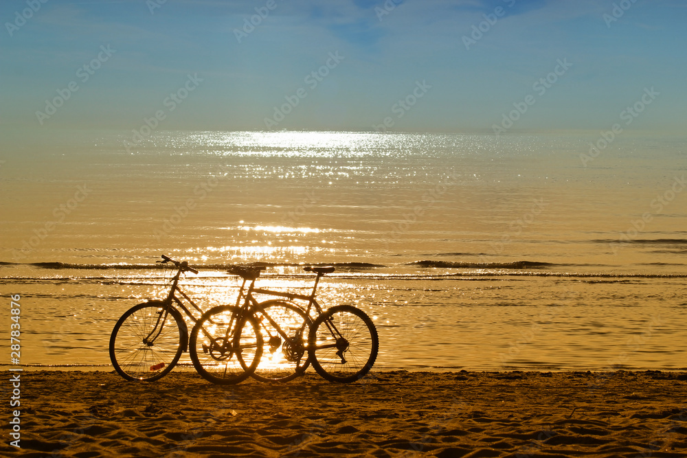 Fototapeta premium Two bicycles silhouettes standing on the sand near the water edge on the beach. Beautiful shiny and glow water of the sea on the background. Sport and recreation concept.