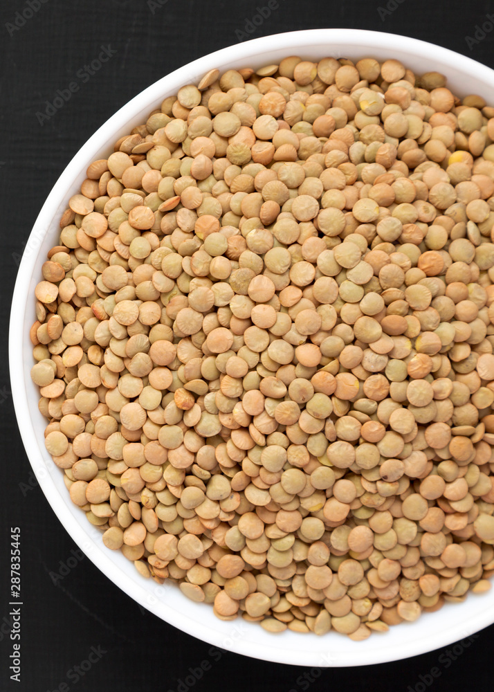 Organic green lentils in a white bowl on a black surface, top view. Flat lay, overhead, from above. Close-up.