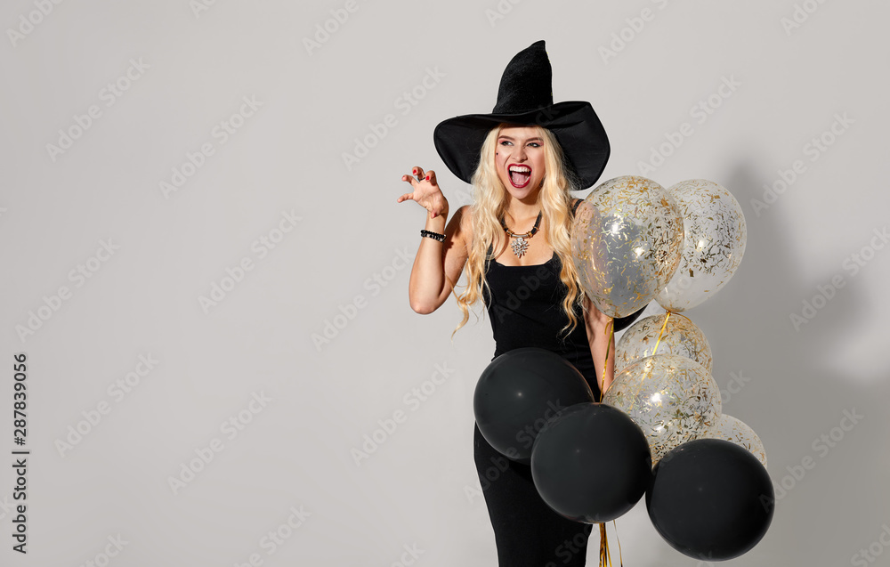 Halloween Party girl. Happy Halloween Sexy Witch with Air balloons. Beautiful young surprised woman in witches hat and short dress holding black balloons. Isolated on gray background.
