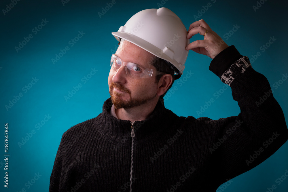 Engineer with protection glasses on scratching the white hardhat helmet. Studio shot of professional with work cap on.