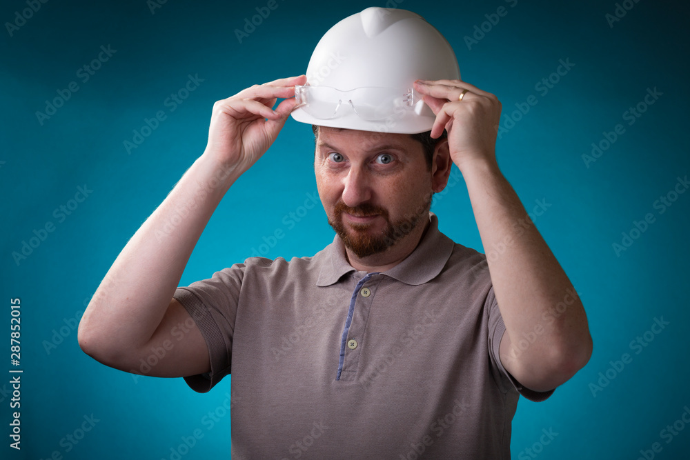 Foreman with beard holding the transparent protective glasses that are on his white safety helmet looking playful at the camera against a studio background and blue spotlight behind Foreman with beard holding the transparent protective glasses that are on his white safety helmet looking playful at the camera against a studio background and blue spotlight behind