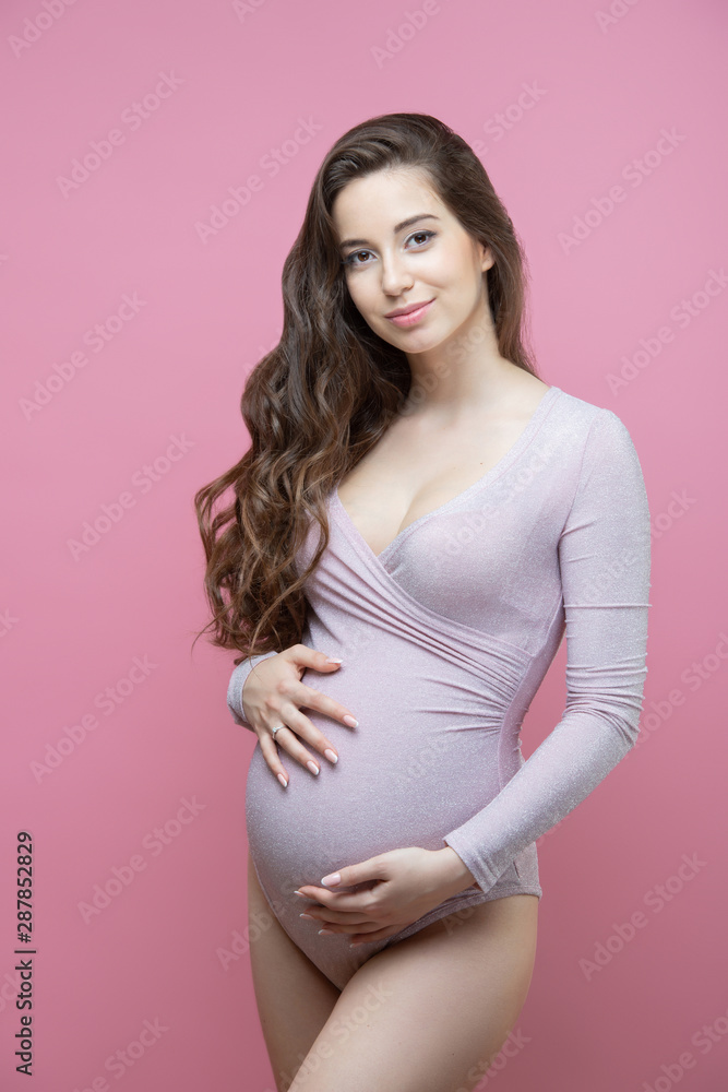 Beautiful young pregnant woman with flowing long wavy hair stands on an isolated pink background and hugs her tummy Beautiful young pregnant woman with flowing long wavy hair stands on an isolated pink background and hugs her tummy