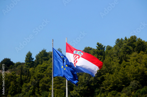 Flags of Croatia and European Union, blowing in the wind. Pine trees and bright sky in the background.