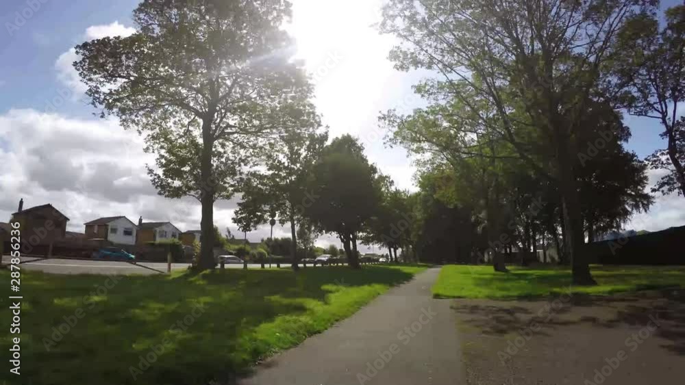 Sunlit park pathway alongside residential town road. Speedy timelapse traffic moving along behind view of trees. Sunlit park pathway alongside residential town road. Speedy timelapse traffic moving along behind view of trees.