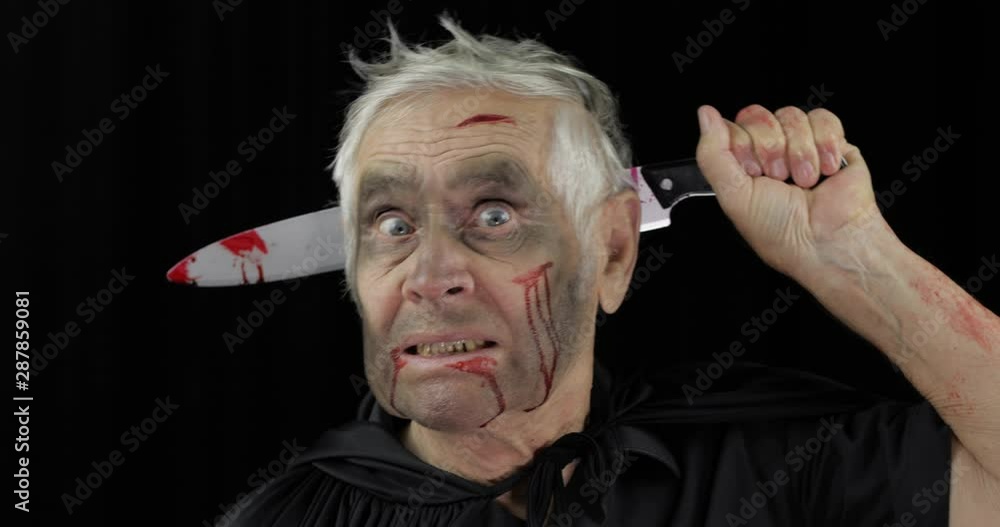 Elderly man with knife in head. Halloween makeup and costume. Blood on his face Elderly man with knife in head. Halloween makeup and costume. Blood on his face