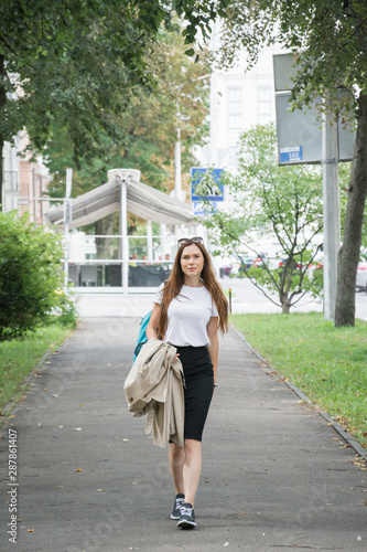 Cheerful attractive young woman with red backpack and book walking in the city