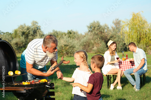 Grandfather with little kids cooking food on barbecue grill and their family in park