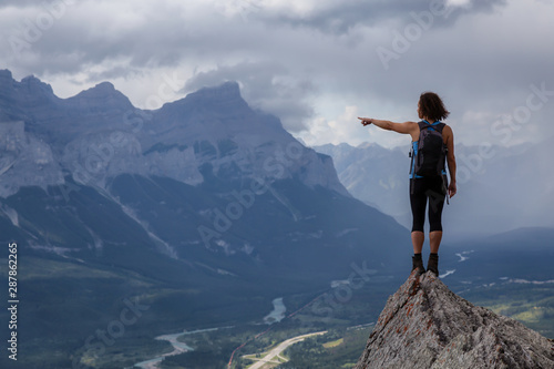 Adventurous Caucasian Girl pointing a finger on top of a rocky mountain during a cloudy and rainy day. Taken from Mt Lady MacDonald, Canmore, Alberta, Canada. © edb3_16