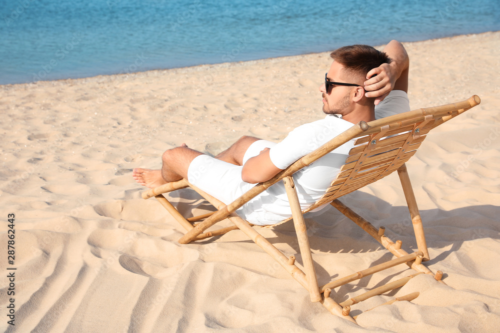 Young man relaxing in deck chair on sandy beach - obrazy, fototapety, plakaty 