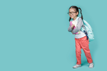 Beautiful smiling Asian little girl with glasses and hold a books with school bag is back to school, empty space in studio shot isolated on colorful blue background, Educational concept for school