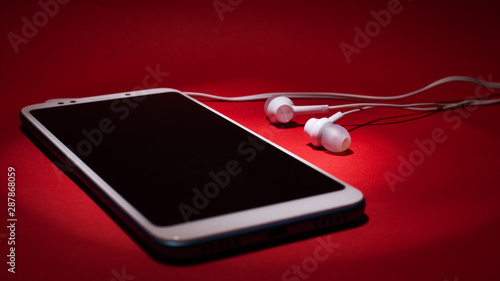 Concept: listen to music from the phone. Smartphone with headphones in the rays of lighton a red background photo