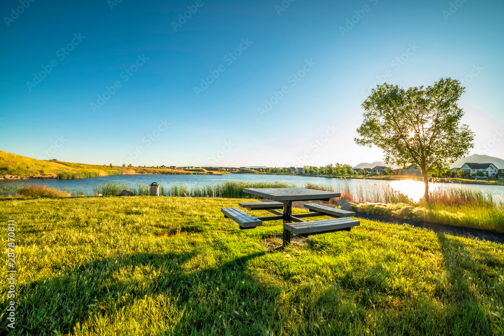 Picnic table at a park with scenic view of a lake that reflects the sky and sun - obrazy, fototapety, plakaty 