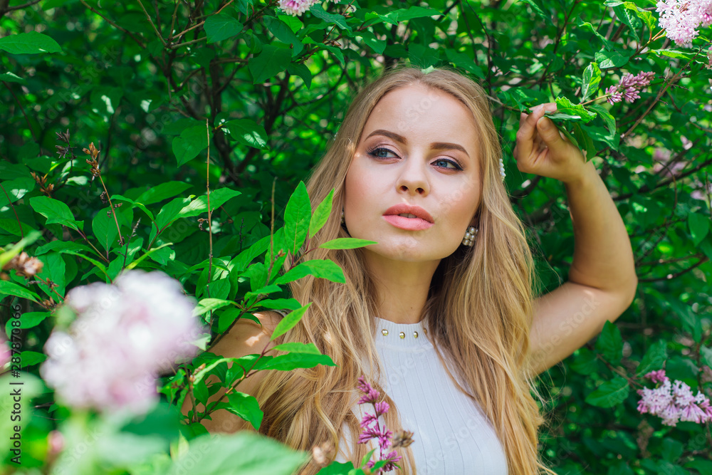 Portrait of a charming blond woman wearing beautiful white dress standing next to lilac bush. Portrait of a charming blond woman wearing beautiful white dress standing next to lilac bush.