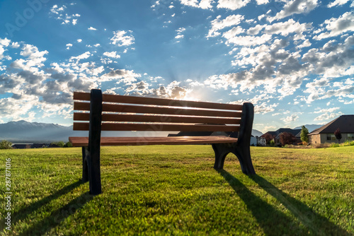 Back view of an outdoor park bench against bright sun and cloudy blue sky