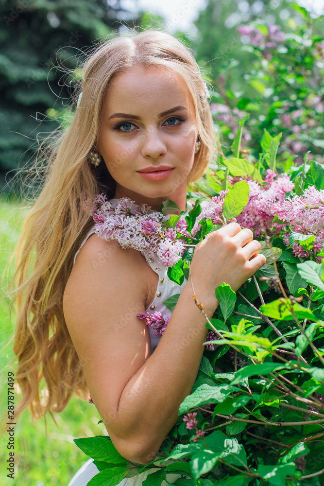 Portrait of a charming blond woman wearing beautiful white dress standing next to lilac bush. Portrait of a charming blond woman wearing beautiful white dress standing next to lilac bush.