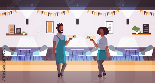 waiters couple carrying coffee and cake on tray african american woman man cafe workers barista in aprons holding cups of cappuccino with desserts modern cafeteria interior full length horizontal © mast3r