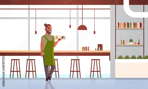 waiter carrying coffee and cake on tray man cafe worker barista in apron holding two cups of cappuccino with dessert modern cafeteria interior full length horizontal