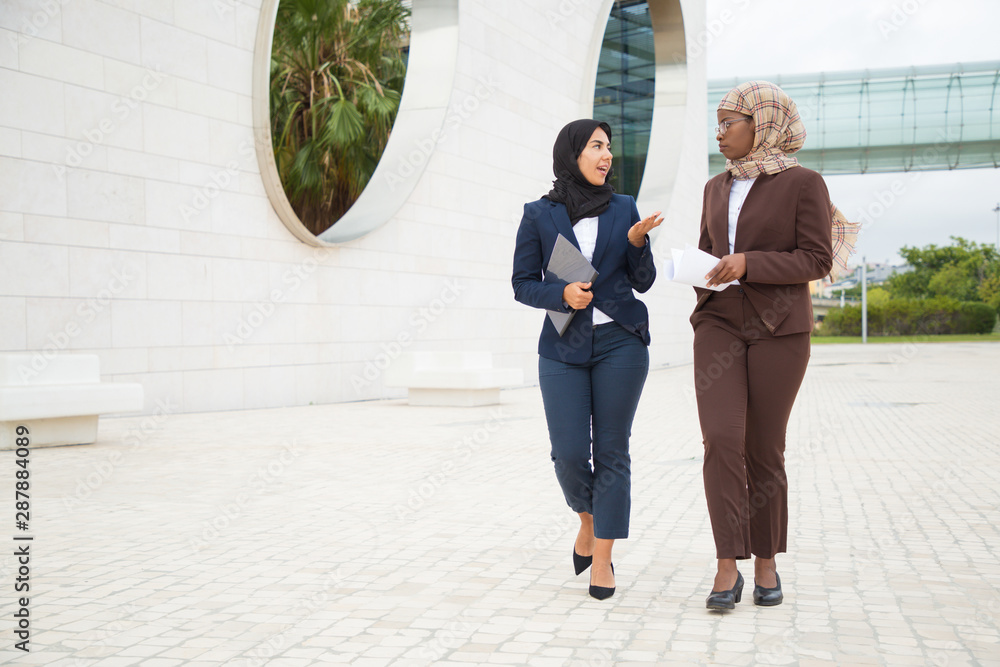 Successful businesswomen walking outside and talking. Muslim women in hijabs and office suits discussing project outdoors. Colleagues concept