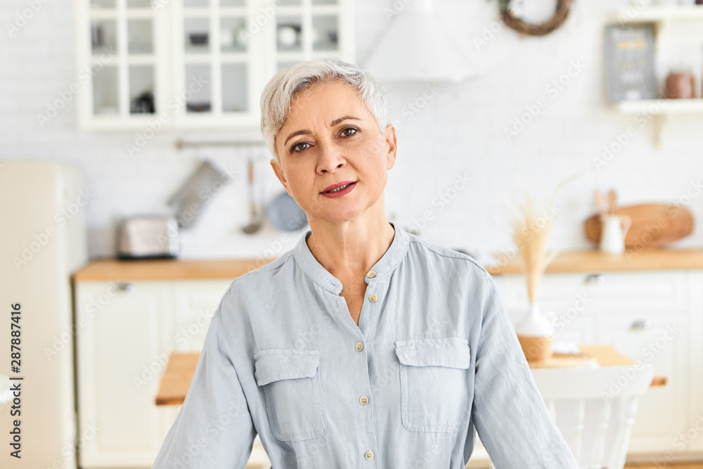 Age, maturity, domesticity and lifestyle. Indoor shot of casually dressed female pensioner having tired look, exhaling after doing all housework, posing against blurred kitchen interior background Age, maturity, domesticity and lifestyle. Indoor shot of casually dressed female pensioner having tired look, exhaling after doing all housework, posing against blurred kitchen interior background
