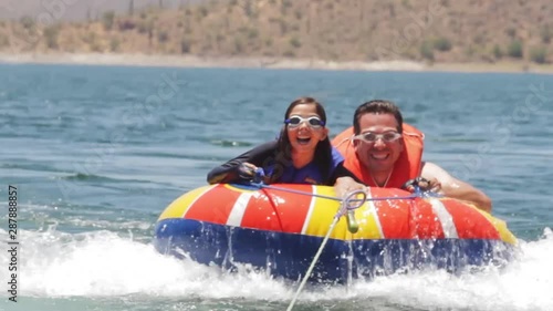 A father and daughter ride on a brightly colored inner tube behind a boat on a desert lake they are wearing goggles and smiling