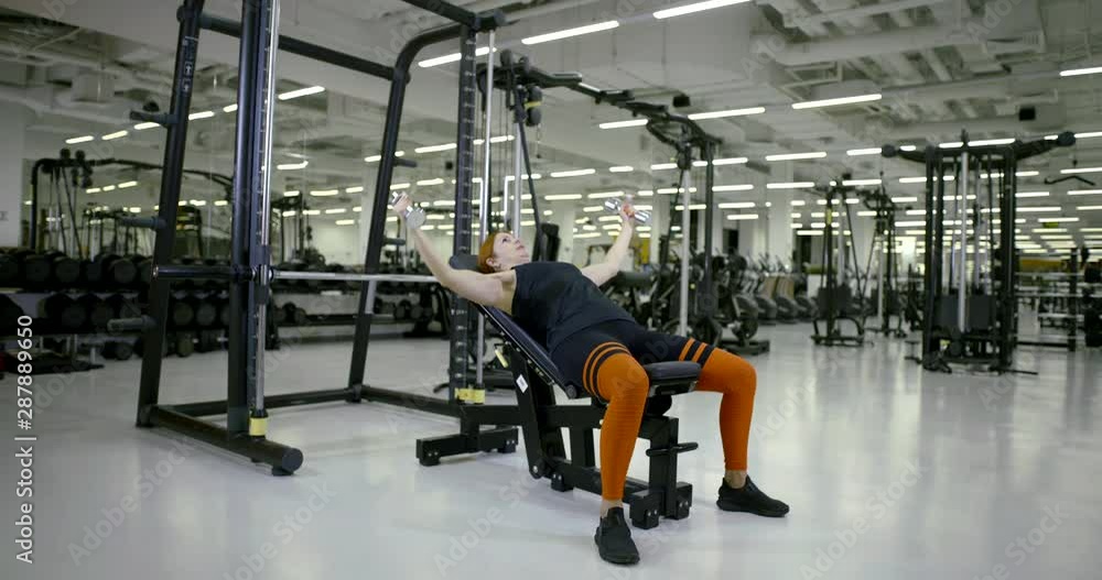 woman is working muscles of chest and hands, lifting dumbbells, lying on sport equipment woman is working muscles of chest and hands, lifting dumbbells, lying on sport equipment