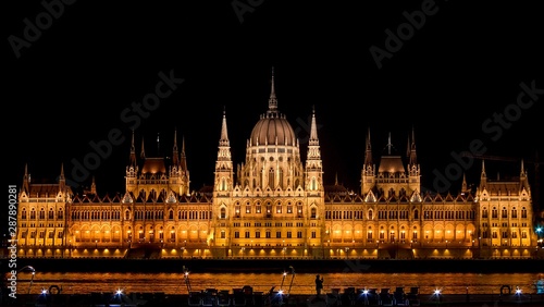Panoramic background orange night image of Budapest parlament from the front with boat and tourist taking a photo of the city.