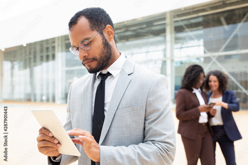 Focused male business professional using tablet outside office building. His female business colleagues using mobile phones behind him. Working outside concept Focused male business professional using tablet outside office building. His female business colleagues using mobile phones behind him. Working outside concept
