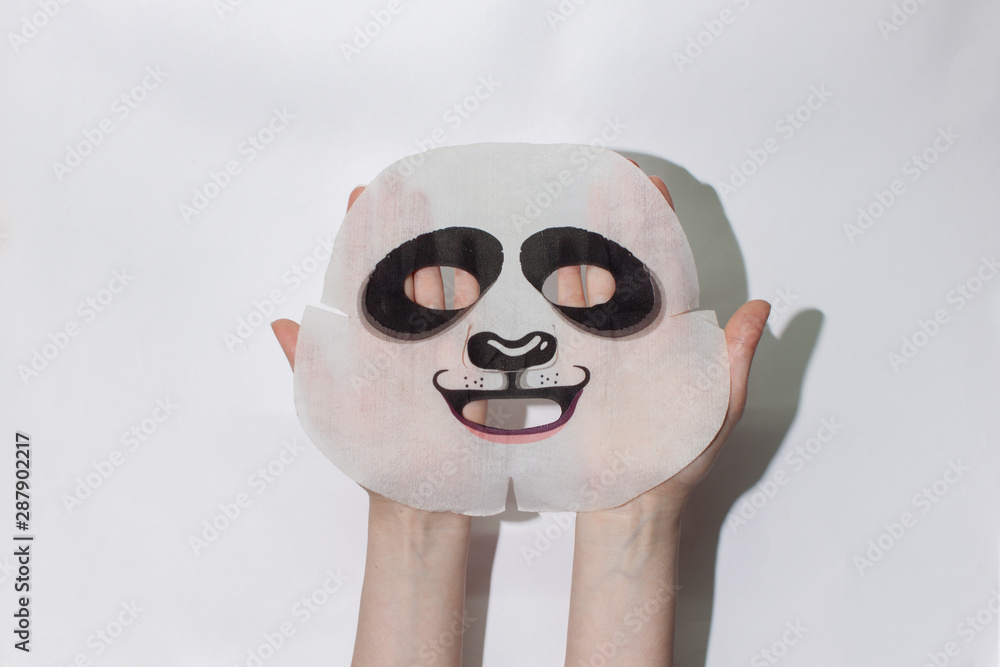 Sheet face mask. Fabric mask for facial skin. Female hands hold a fabric panda  mask for the face. On a white background. View from above. Cosmetology,  medicine and healthcare. Stock Photo
