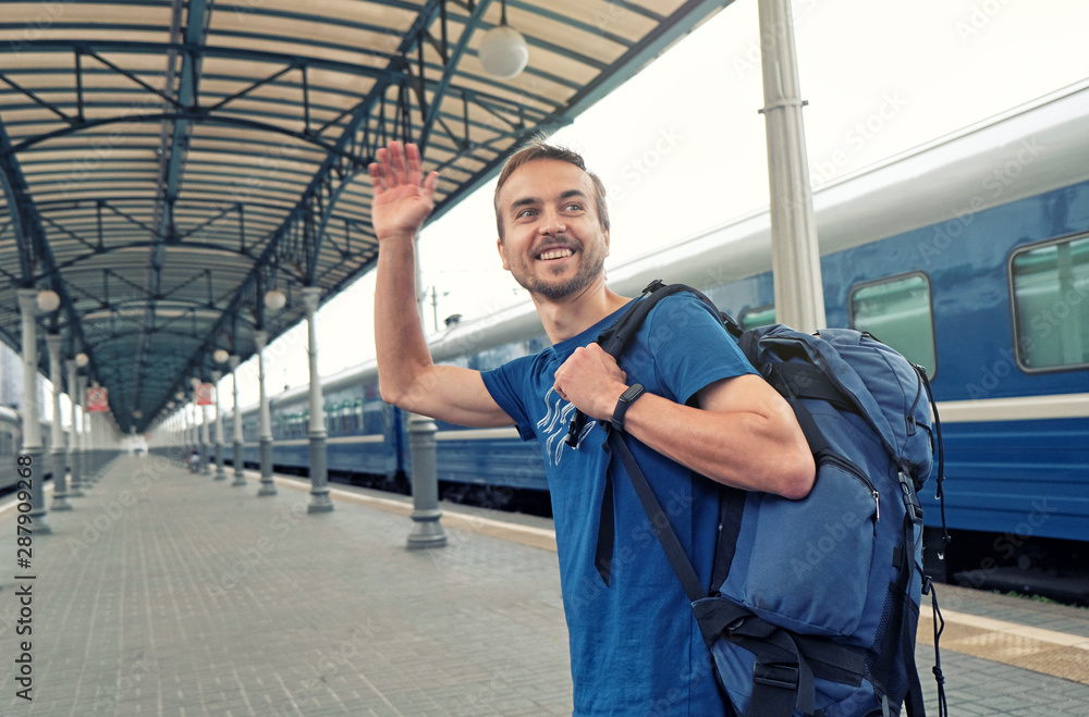 Happy man tourist with backpack stand on railway station platform, greeting friends or saying goodbye, waving his hand. Travel by train. Happy man tourist with backpack stand on railway station platform, greeting friends or saying goodbye, waving his hand. Travel by train.