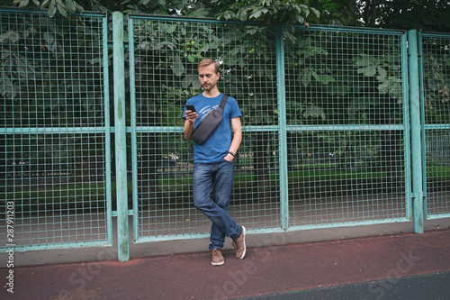 Urban lifestyle. Young bearded man with smartphone and crossbody bag staying near fence of city sport playground. Copy space. photo