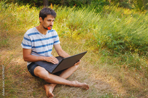 A young man sits on the grass with a flower in his mouth and works correspondence on a laptop.