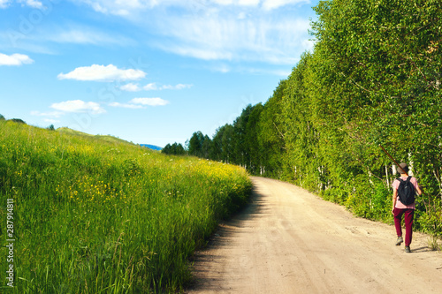 A Guy in a Pink T-Shirt, Red Pants, and Bucket Hat, with Black Backpack and Long Pole on His Shoulder Walks along Highland Country Road. Sunny Spots on Grassy Hillside and Birch Trees along the Road.