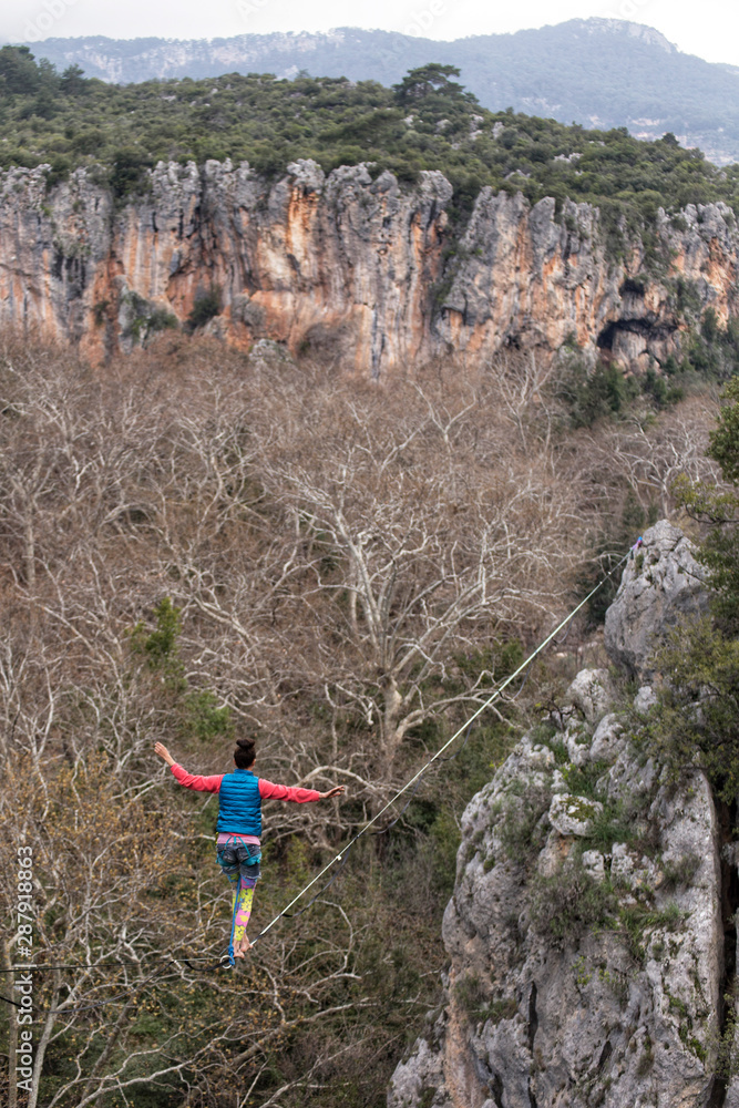 Fototapeta premium A woman is walking along a stretched sling. Highline in the mountains. Woman catches balance. Performance of a tightrope walker in nature. Highliner on the background of valley.