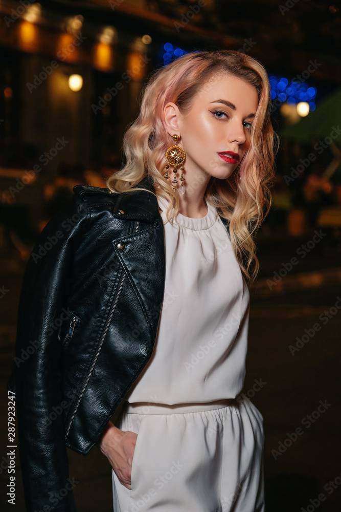Luxury beautiful blond woman with curly hair and evening make up standing in the middle of night city. She wears casual white blouse and trousers and leather jacket Luxury beautiful blond woman with curly hair and evening make up standing in the middle of night city. She wears casual white blouse and trousers and leather jacket