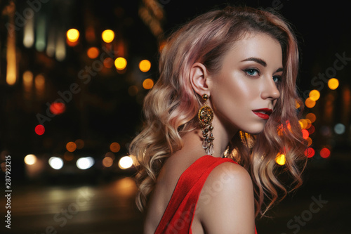 Luxury beautiful blond woman with curly hair and evening make up standing in the middle of night city. She wears red romantic dress