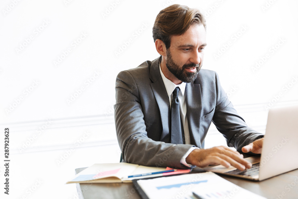 Handsome young positive business man sitting in office using laptop computer working indoors. Handsome young positive business man sitting in office using laptop computer working indoors.