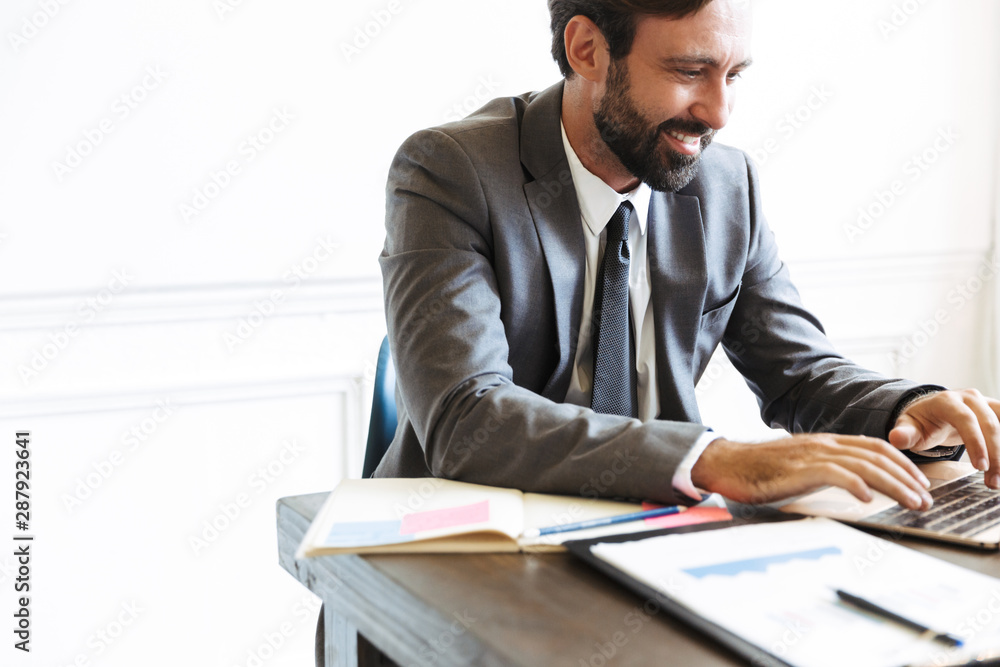 Handsome young positive business man sitting in office using laptop computer working indoors. Handsome young positive business man sitting in office using laptop computer working indoors.