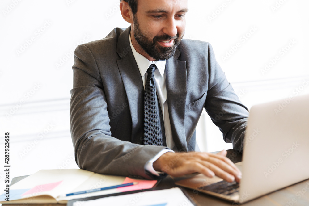 Handsome young positive business man sitting in office using laptop computer working indoors. Handsome young positive business man sitting in office using laptop computer working indoors.