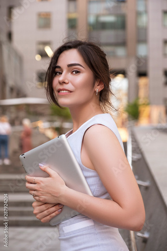 Beautiful brunette business woman in grey smark casual dress working on a tablet in her hands outdoors. European city on background. copy space