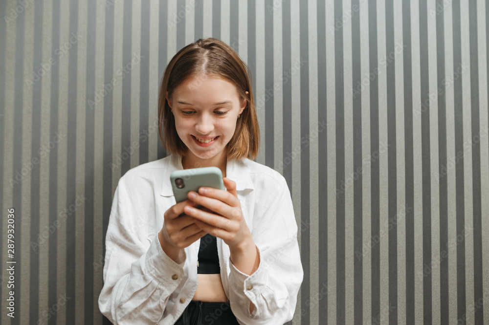 Portrait of happy girl with smartphone in hands on gray wall background, uses phone, looks at screen and smiling, wears white shirt. Smiling stylish girl communicates on social networks. Portrait of happy girl with smartphone in hands on gray wall background, uses phone, looks at screen and smiling, wears white shirt. Smiling stylish girl communicates on social networks.