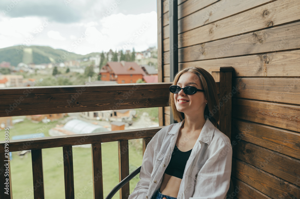 Happy girl in sunglasses sits on the balcony in the cottage,smiling woman satisfied with vacation in hotel.Smiling stylish girl sitting on the balcony of the hotel overlooking the mountains and houses