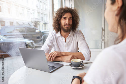 Young good looking bearded businessman having appointment out of office  having pleasant talk in cafe while drinking coffee  wearing white shirt
