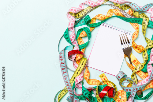 Top view of notebook with fork surrounded with colorful measuring tapes on blue background. Copy space with diet planning